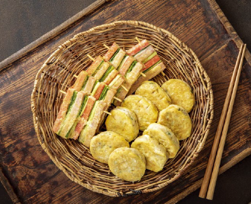 Wicker basket with assorted Korean jeon pancakes on a wooden tray. Includes colorful skewered vegetable and ham slices. Mood: appetizing.