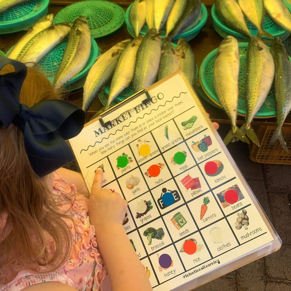 Child holds a bingo card at a fish market, pointing at items. Fresh fish on green baskets in the background. Bright, playful mood.