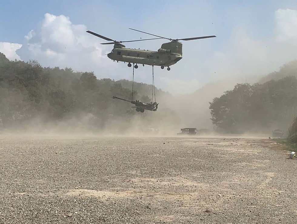 A helicopter lifts artillery on a dusty field with forested hills in the background. Dust is swirling, creating an intense and dynamic scene.