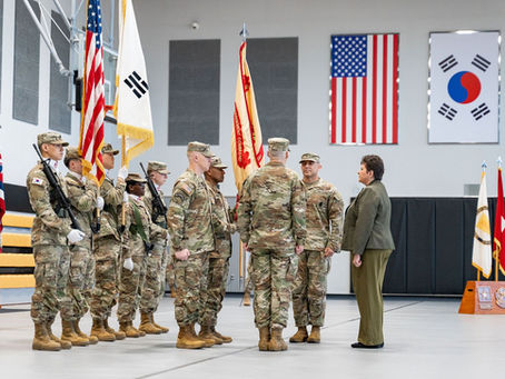 Soldiers in camouflage uniforms stand in formation holding flags; U.S. and South Korean flags hang on the wall. A woman in a suit faces them.