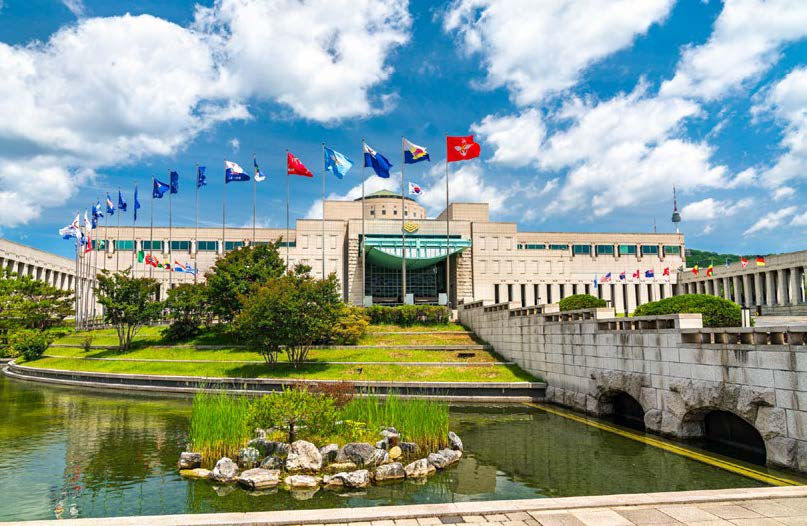 A large building with multiple flags sits under a blue sky. A pond and greenery are in the foreground, creating a serene atmosphere.