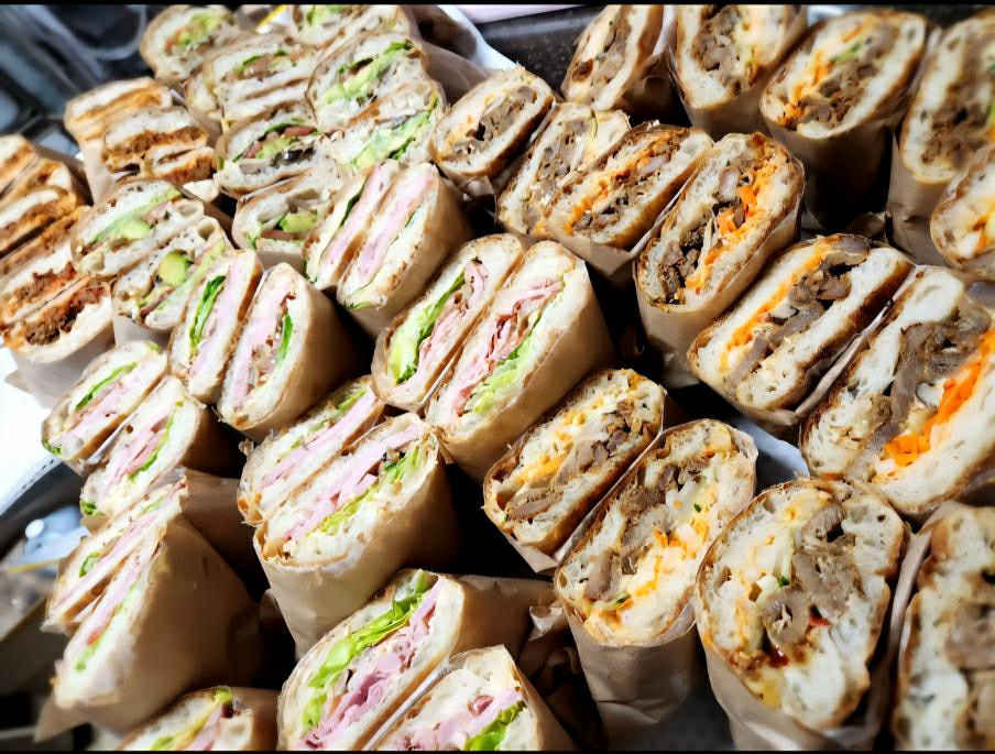 Rows of assorted sandwiches with various fillings like lettuce, ham, and cheese in paper wrappers on a table, evoking a fresh, vibrant mood.