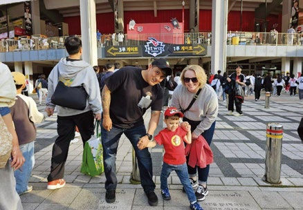 Family of three smiling outdoors near a stadium entrance. The child wears a red cap. Crowd and signage visible in the background.