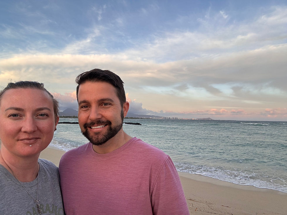 A smiling couple takes a selfie on a sandy beach at sunset, with a calm ocean and cloudy sky in the background.
