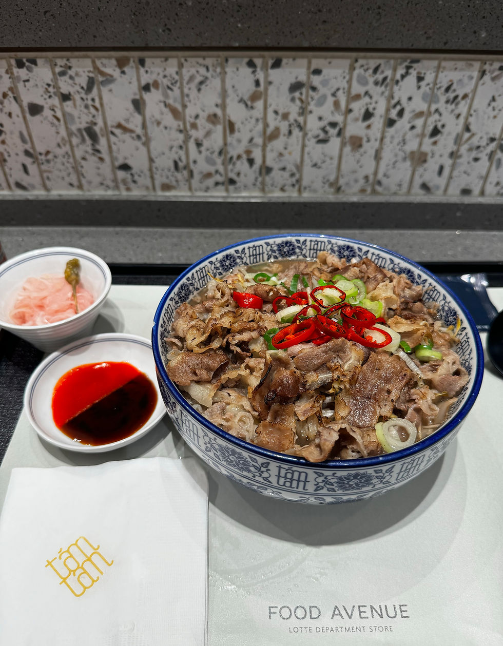 Bowl of beef rice topped with red and green chili, beside soy sauce and pickles. Napkin reads tam tam. Terrazzo background.