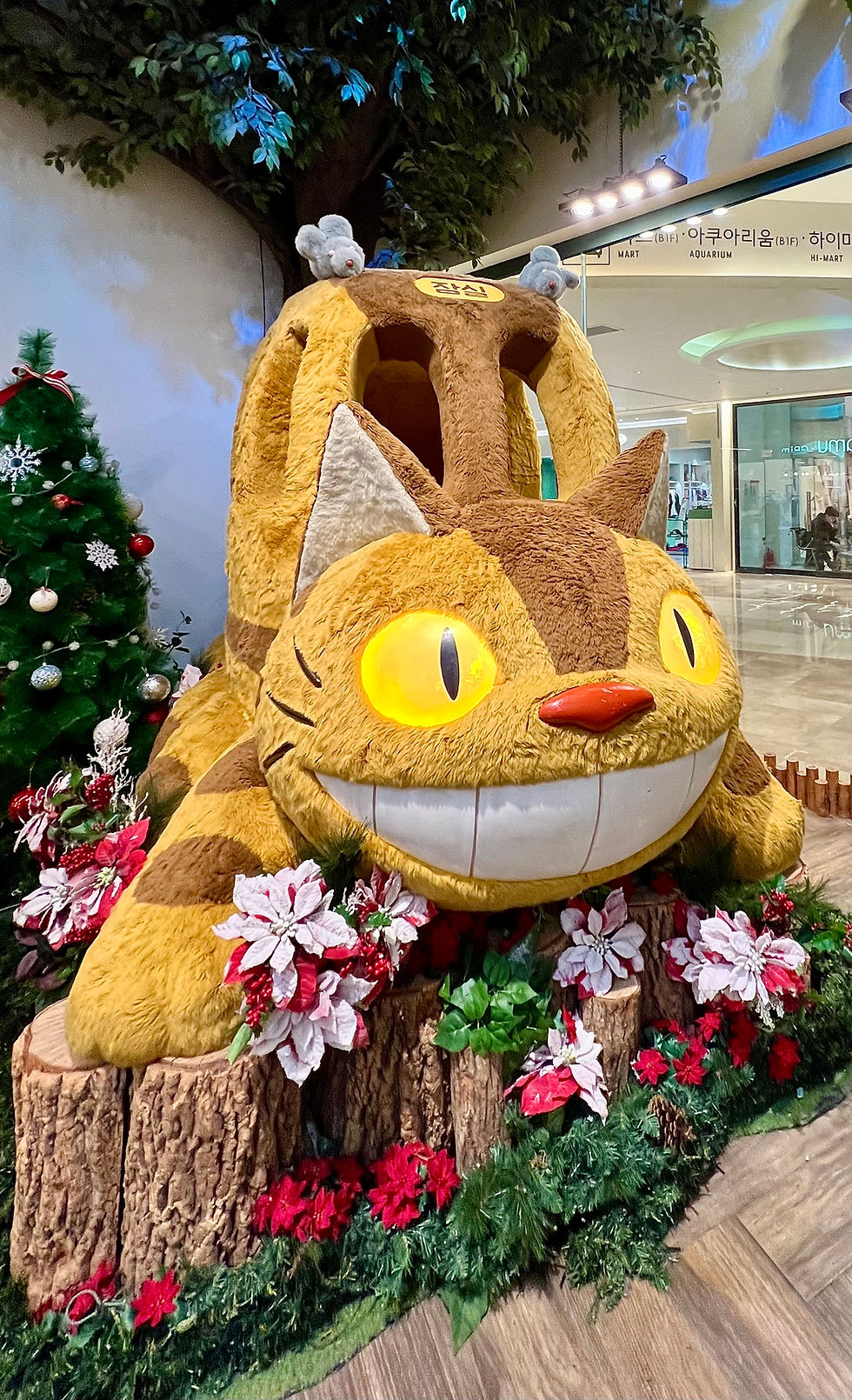 Large plush cat bus with glowing eyes, adorned in holiday decor, including red poinsettias and a nearby Christmas tree, in a mall.
