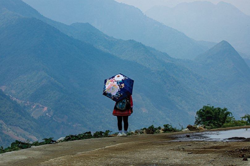 A person with a floral umbrella stands on a road, overlooking misty blue mountains. The scene is peaceful and scenic.