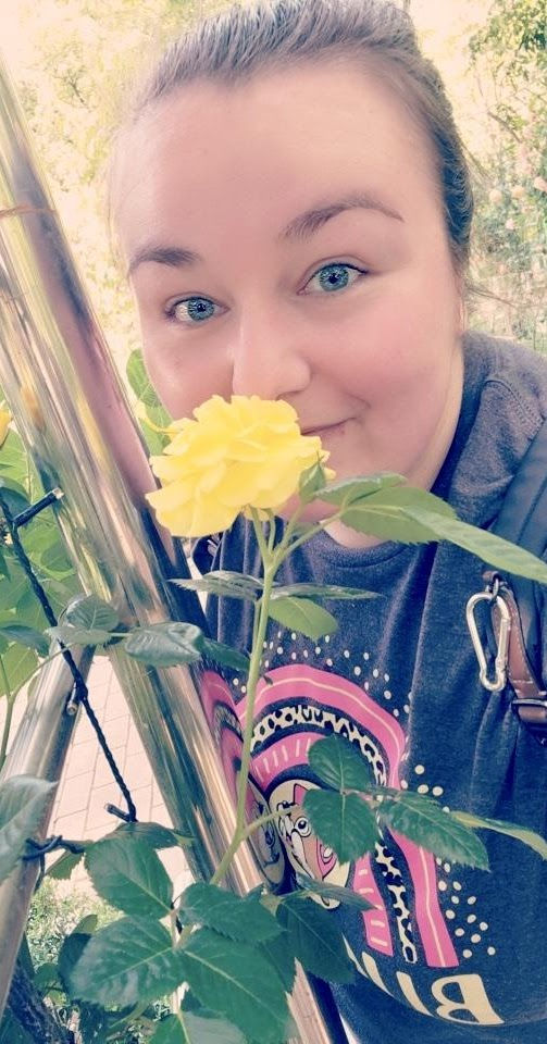 Smiling person leaning near a yellow rose, wearing a colorful shirt and backpack strap; background is bright and leafy.