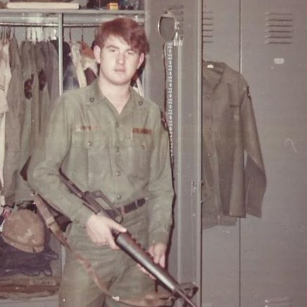 Young man in military uniform holds a rifle in front of open lockers with clothes and gear. Neutral expression, indoor setting.