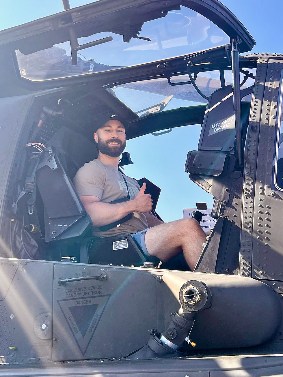 Man smiling in helicopter cockpit, wearing a beige shirt and cap, giving thumbs up. Sunlit interior, "DO NOT GRAB" sign visible.