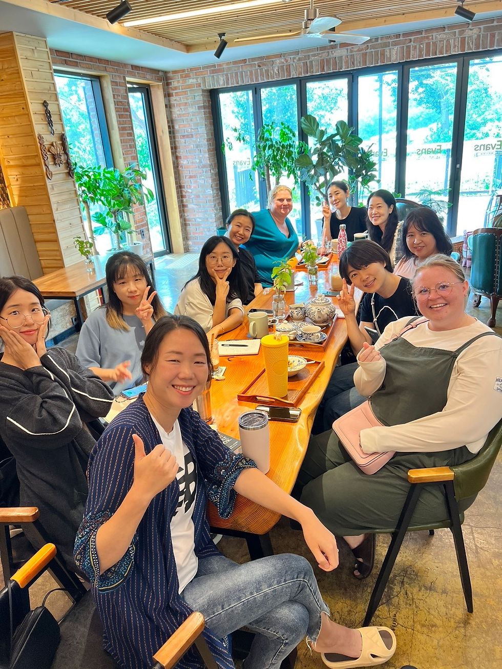 A group of smiling people sits around a wooden table in a cozy cafe with plants and large windows. Some make peace signs, creating a joyful mood.