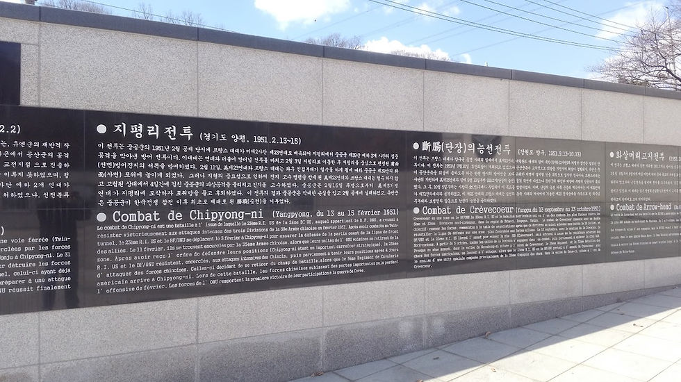 Black plaques on a gray wall display text in Korean and French, detailing historical battles. Sunny day with clear skies and tree shadows.