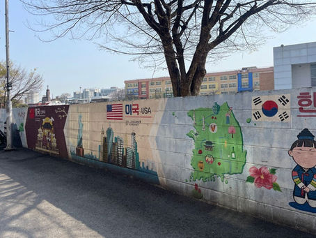 Colorful mural on a wall depicting USA, China, and Korea with maps and symbols. Korean flag and smiling child in traditional attire. School in background.