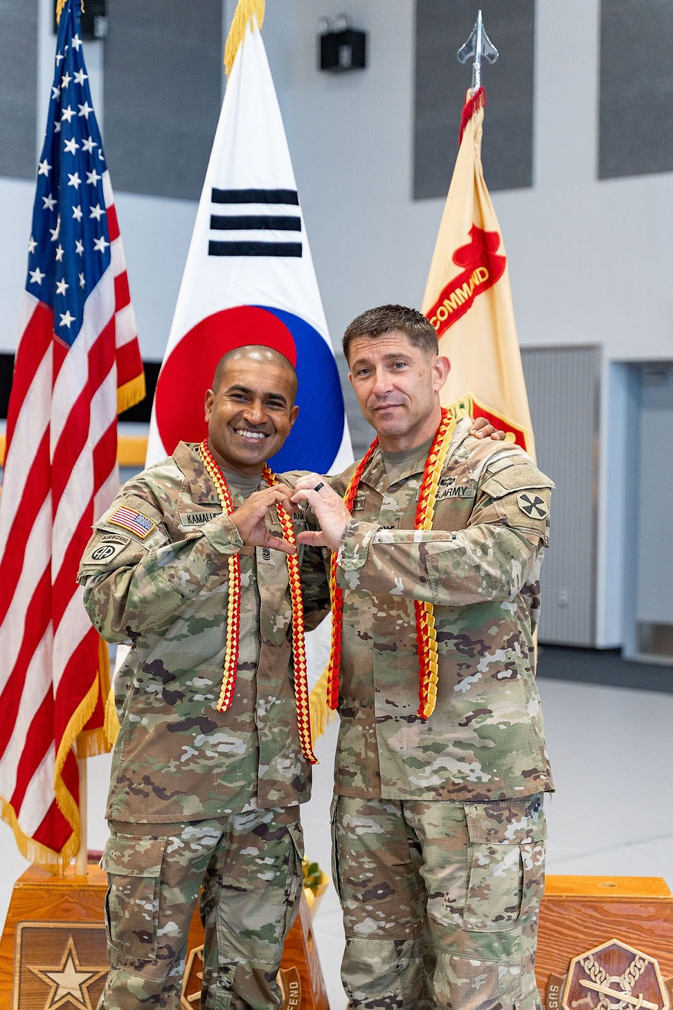 Two soldiers in camo uniforms make a heart shape with hands, smiling. Flags in background are American and Korean. They wear red and yellow leis.