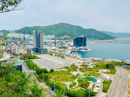 Overlooking a coastal cityscape with modern buildings, winding roads, green parks, and a harbor. Lush greenery and distant mountains.