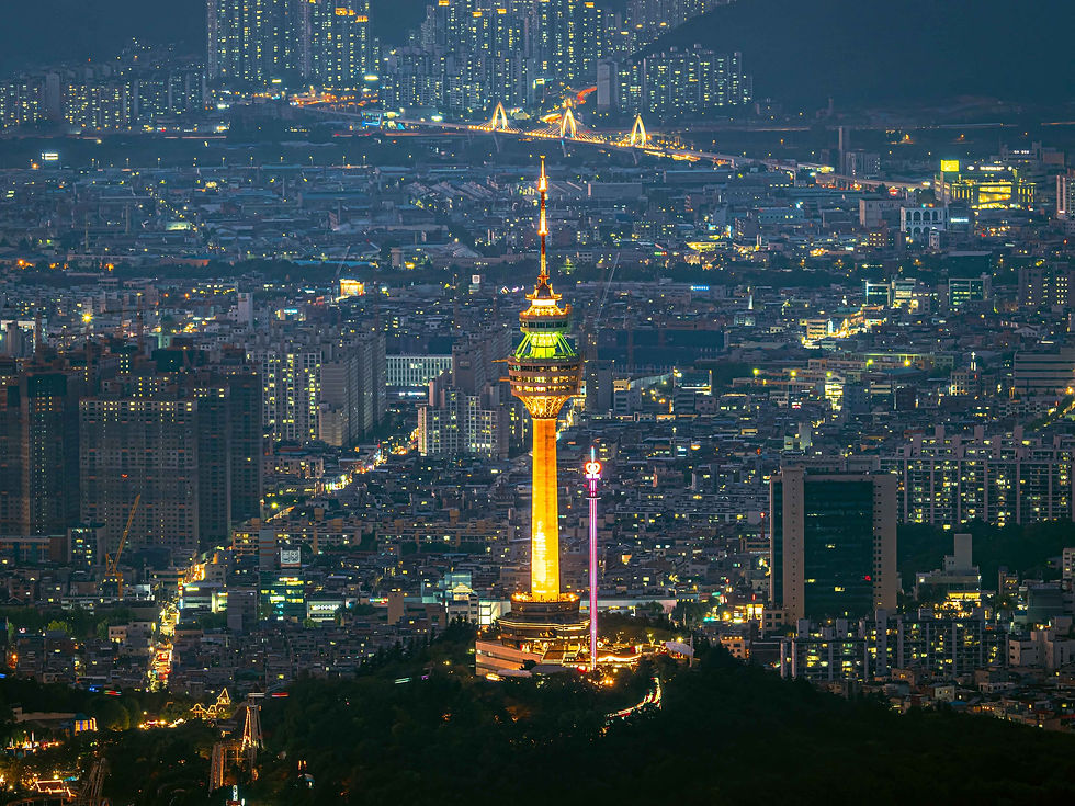 Cityscape at night featuring a tall, illuminated tower in the foreground. Buildings and streets with glowing lights fill the background.