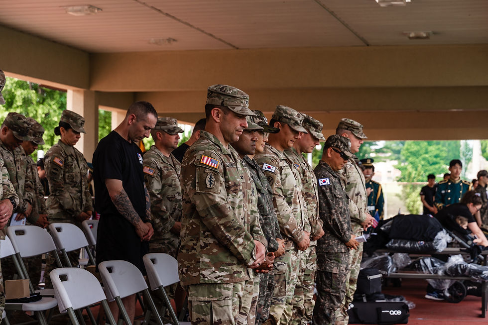 Soldiers in camouflage and one in black stand with heads bowed in a solemn ceremony under a covered area. Korean flags visible on uniforms.