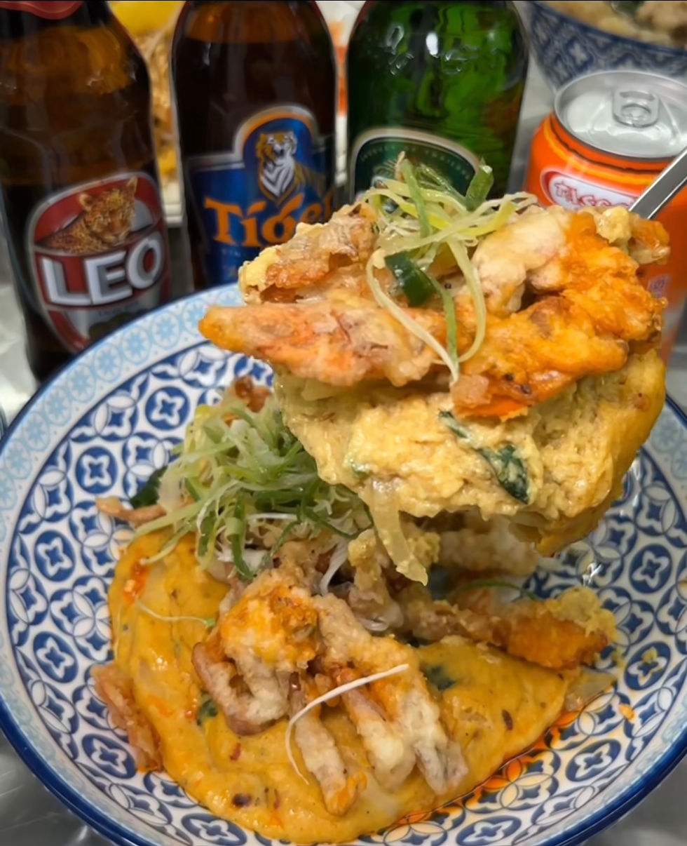 Colorful noodle dish topped with fried seafood and green onions in a patterned bowl. Beer bottles and an orange can are in the background.