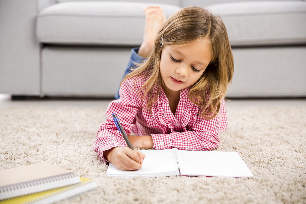Child in red checkered shirt writing in a notebook on a beige carpet, with a couch in the background. Notebooks are scattered nearby.