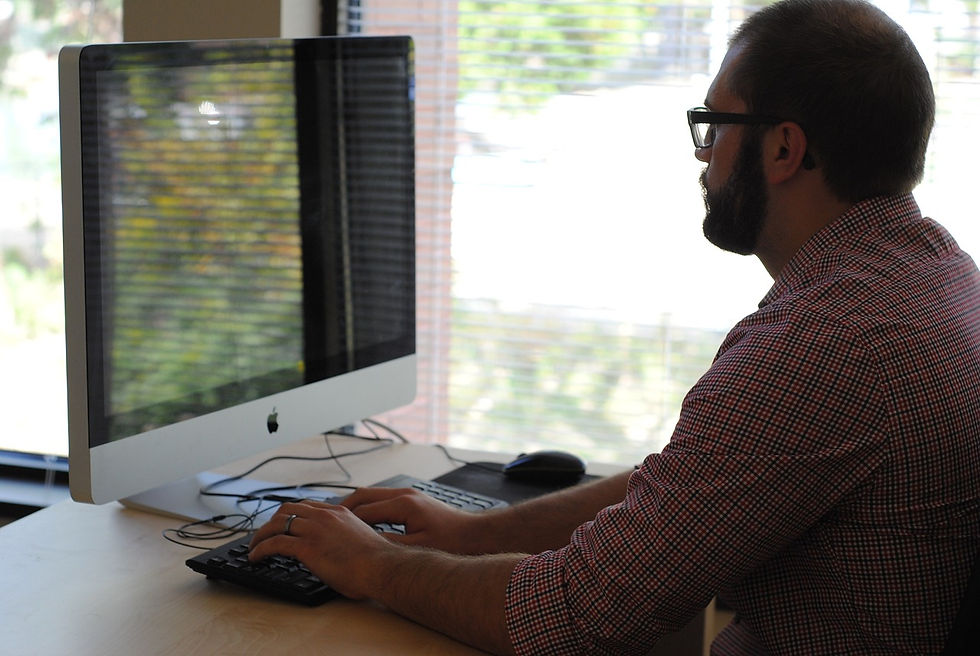 Man in a red checkered shirt types on a computer in a bright office with window blinds. Calm atmosphere, screen shows an Apple logo.