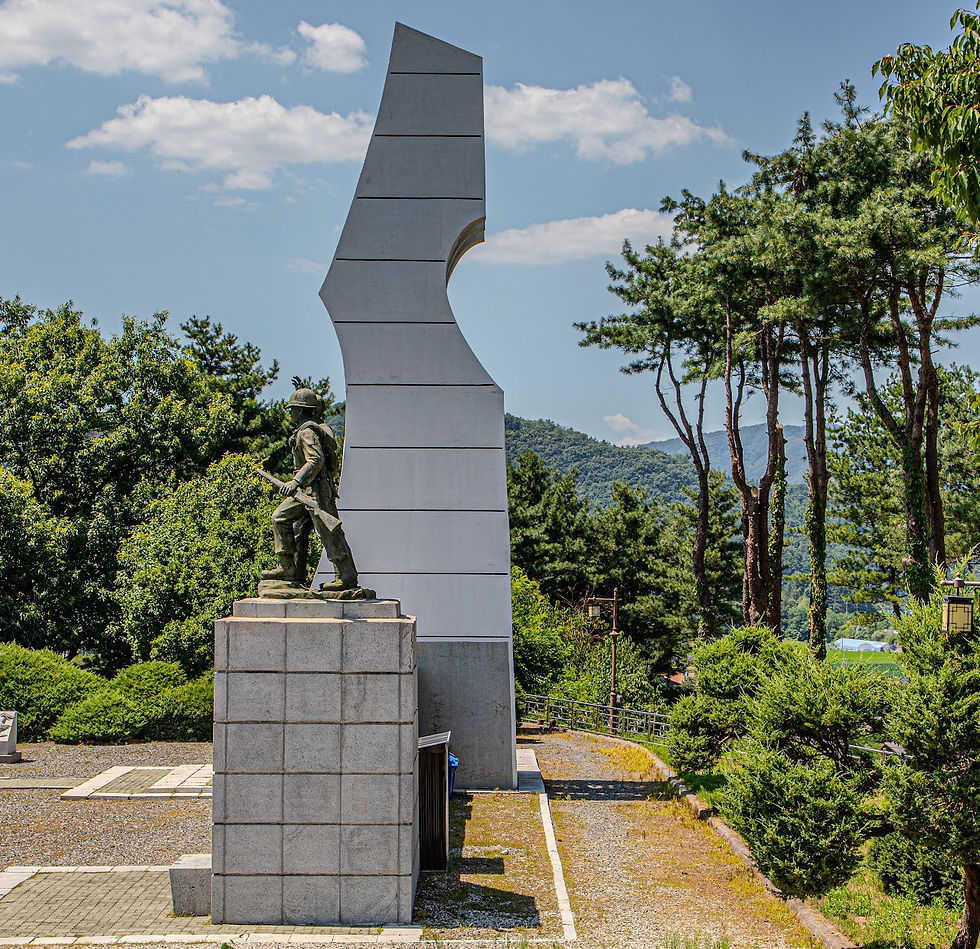 A soldier statue stands on a pedestal next to a modern white monument. Trees and mountains are in the background under a blue sky with clouds.