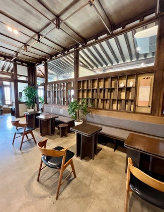 Cozy cafe interior with wooden tables and chairs, plants, and a large mirror reflecting ceiling beams. Warm and inviting atmosphere.