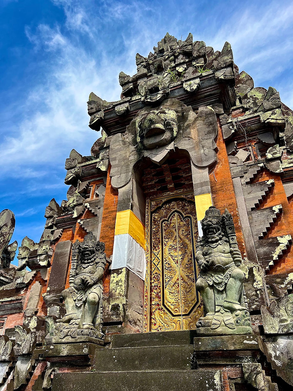 Ornate Balinese temple gate with stone statues on either side, intricate gold door, and vivid blue sky background conveying serenity.