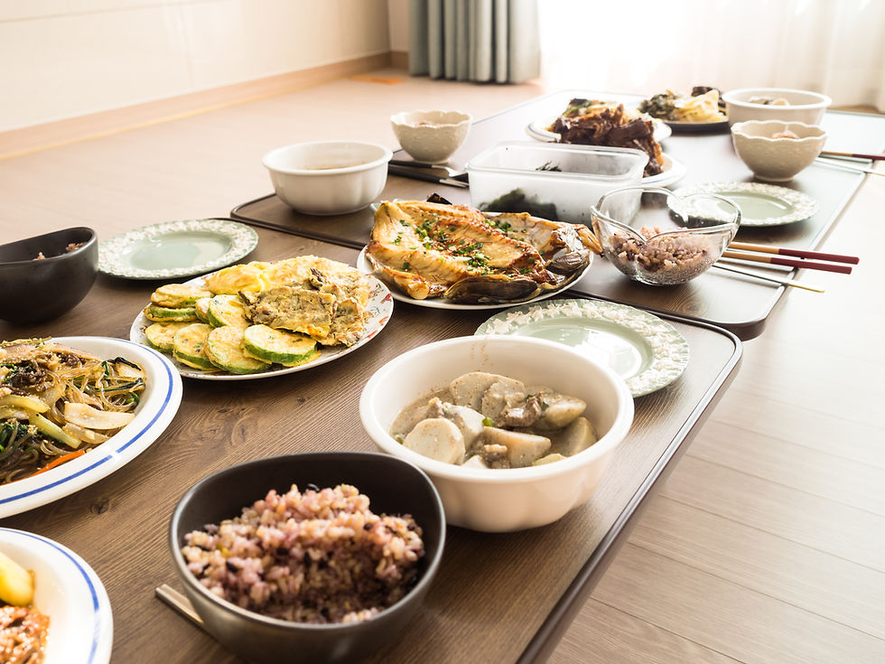 A table with various Korean dishes, including seafood, pancakes, and rice, on a wooden surface. Bright, natural light fills the room.