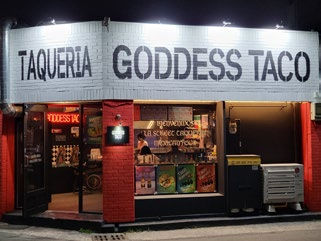 Taco restaurant at night with "Taqueria Goddess Taco" lit sign. Red and white exterior, colorful posters in window, inviting mood.