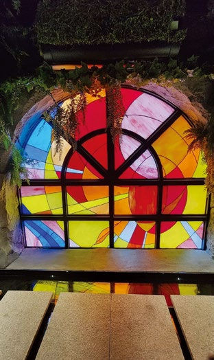 Colorful stained glass window with abstract patterns, set in a lush green background. Reflective water surface in front.
