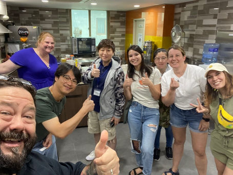 Group of eight people in a kitchen, smiling and giving thumbs up. Bright, colorful setting with a tiled background, conveying a joyful mood.