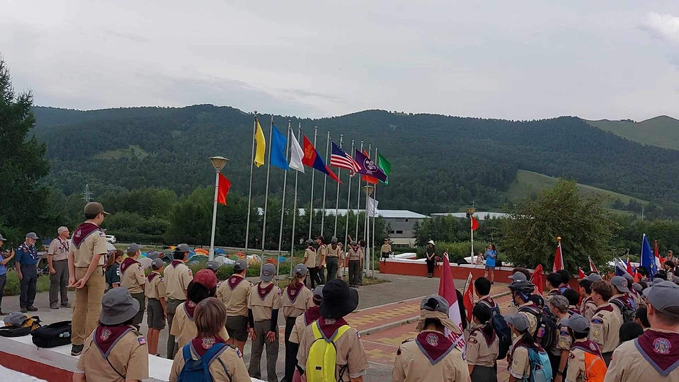 Scouts in uniforms gather near flagpoles with colorful flags against a mountainous backdrop. The atmosphere is organized and spirited.