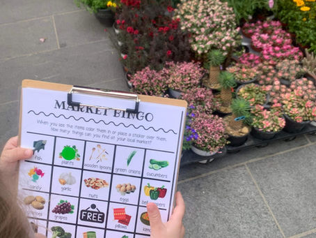 Child holds a "Market Bingo" board beside colorful plants in a market. The bingo sheet features items like fish and plants. Urban setting.