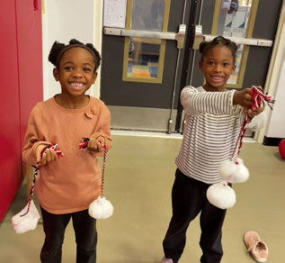 Two children smiling, holding white pompoms on strings, standing in a hallway with a red wall. Shoes on the floor. Joyful mood.