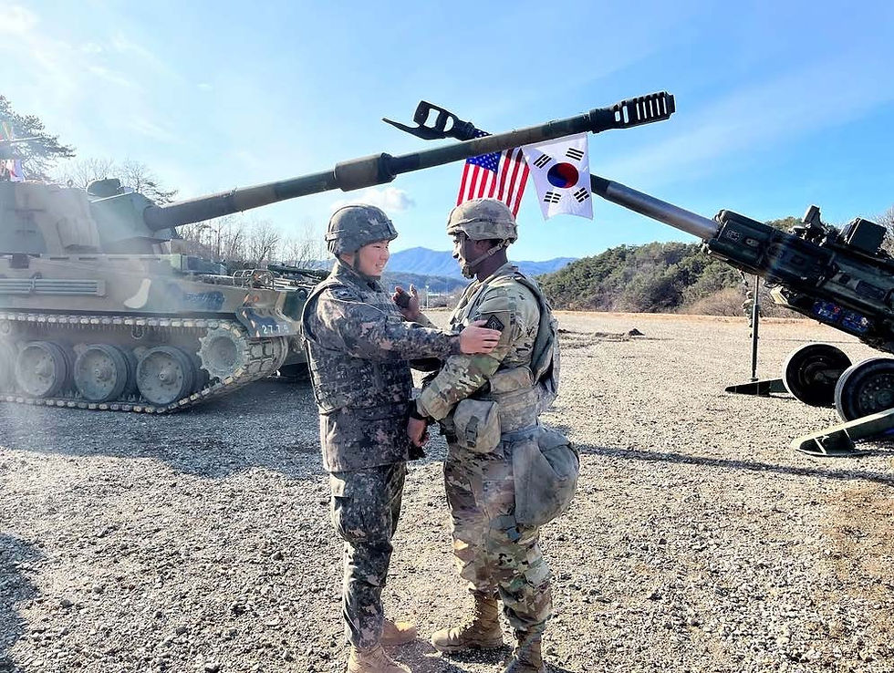 Two soldiers in camouflage uniforms greet each other warmly. Tanks in the background with U.S. and South Korean flags. Blue sky.