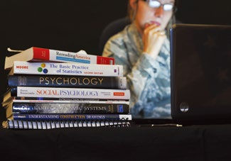 A person in glasses, deep in thought, looks at a laptop. A stack of academic books, including "Psychology" and "Statistics," is beside them.