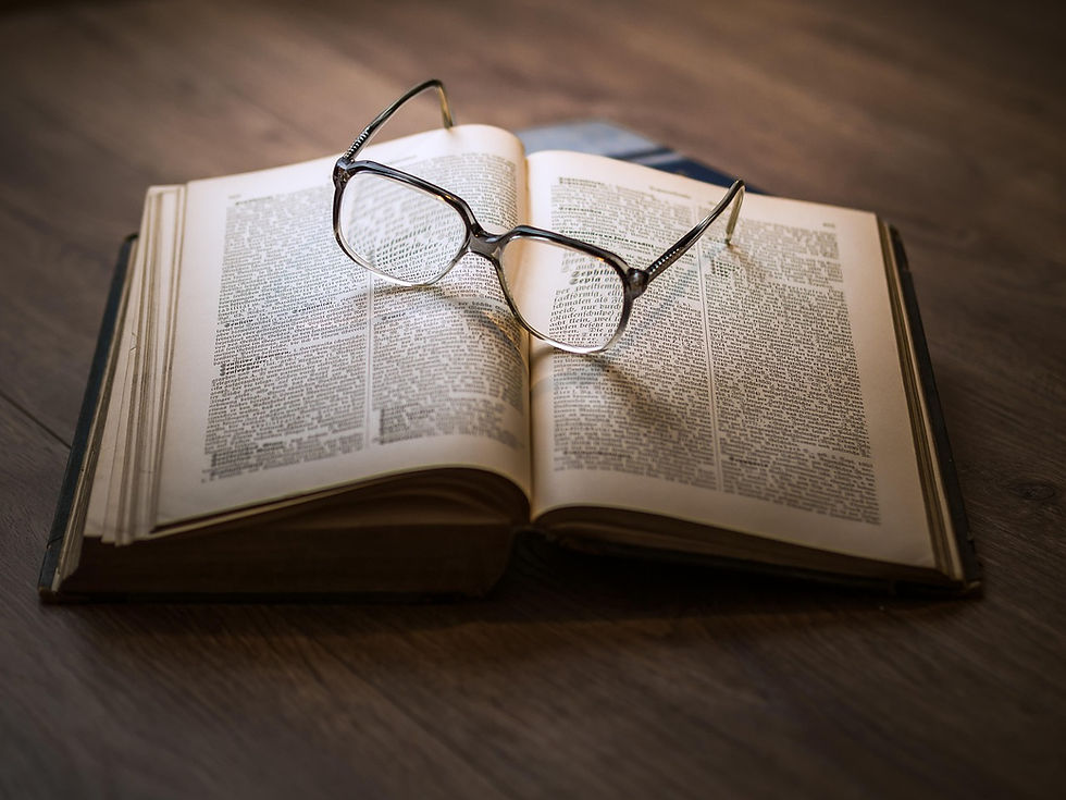 Open book with glasses on top, resting on a wooden surface. Pages have small text, creating a studious and serene atmosphere.