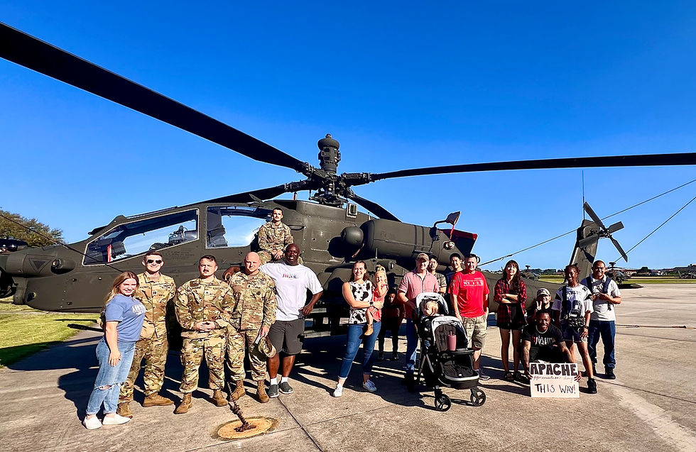Group of people, including military personnel, pose in front of an Apache helicopter on a sunny day. One holds a "Apache This Way" sign.