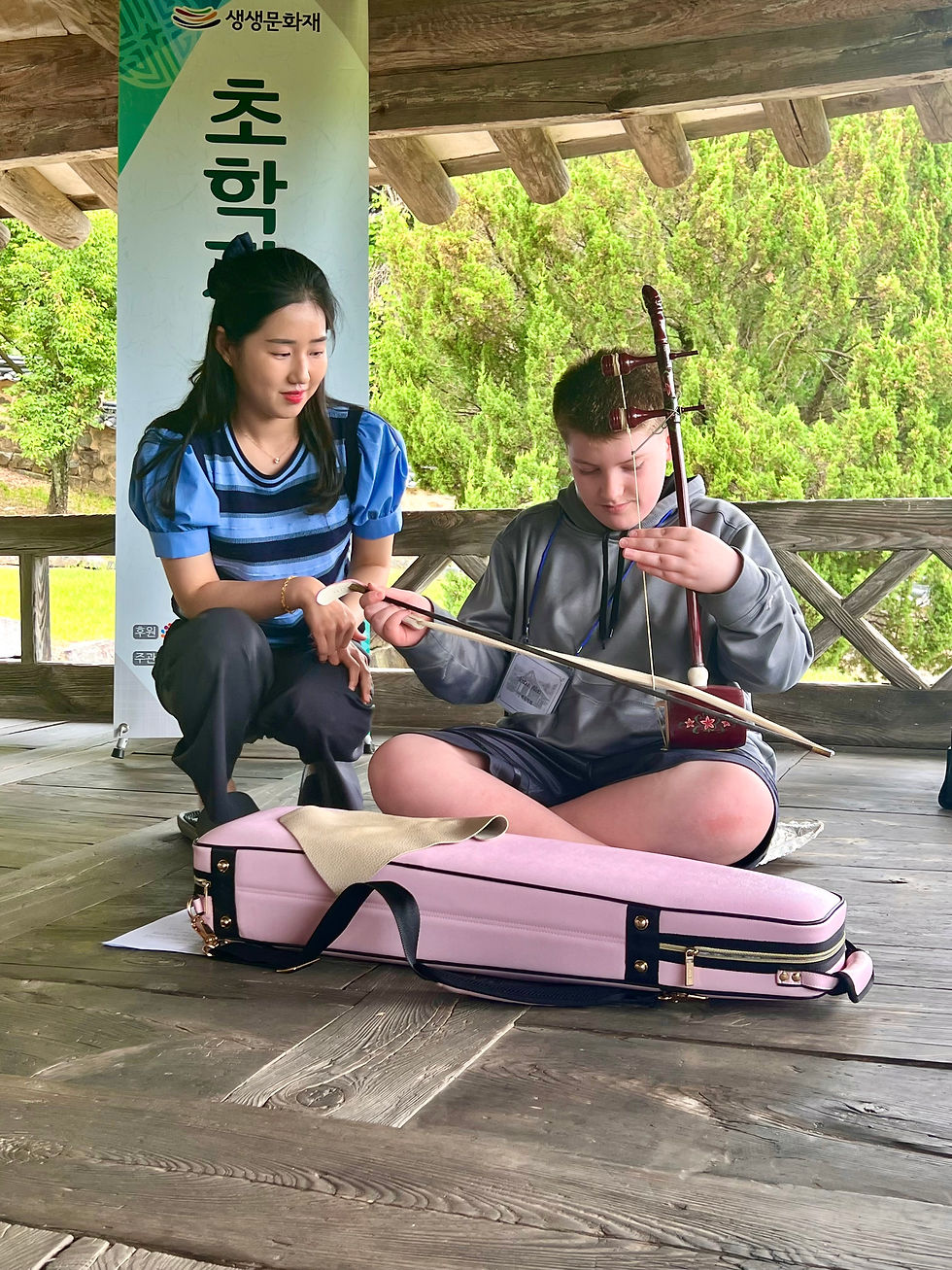 A woman teaches a boy to play a traditional string instrument on a wooden porch, with a pink case nearby. Lush greenery in the background.