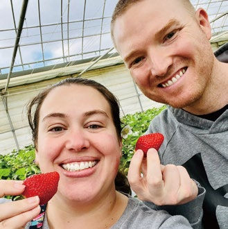 Smiling man and woman hold strawberries in a greenhouse, surrounded by green plants. They appear happy. The man wears a gray hoodie.