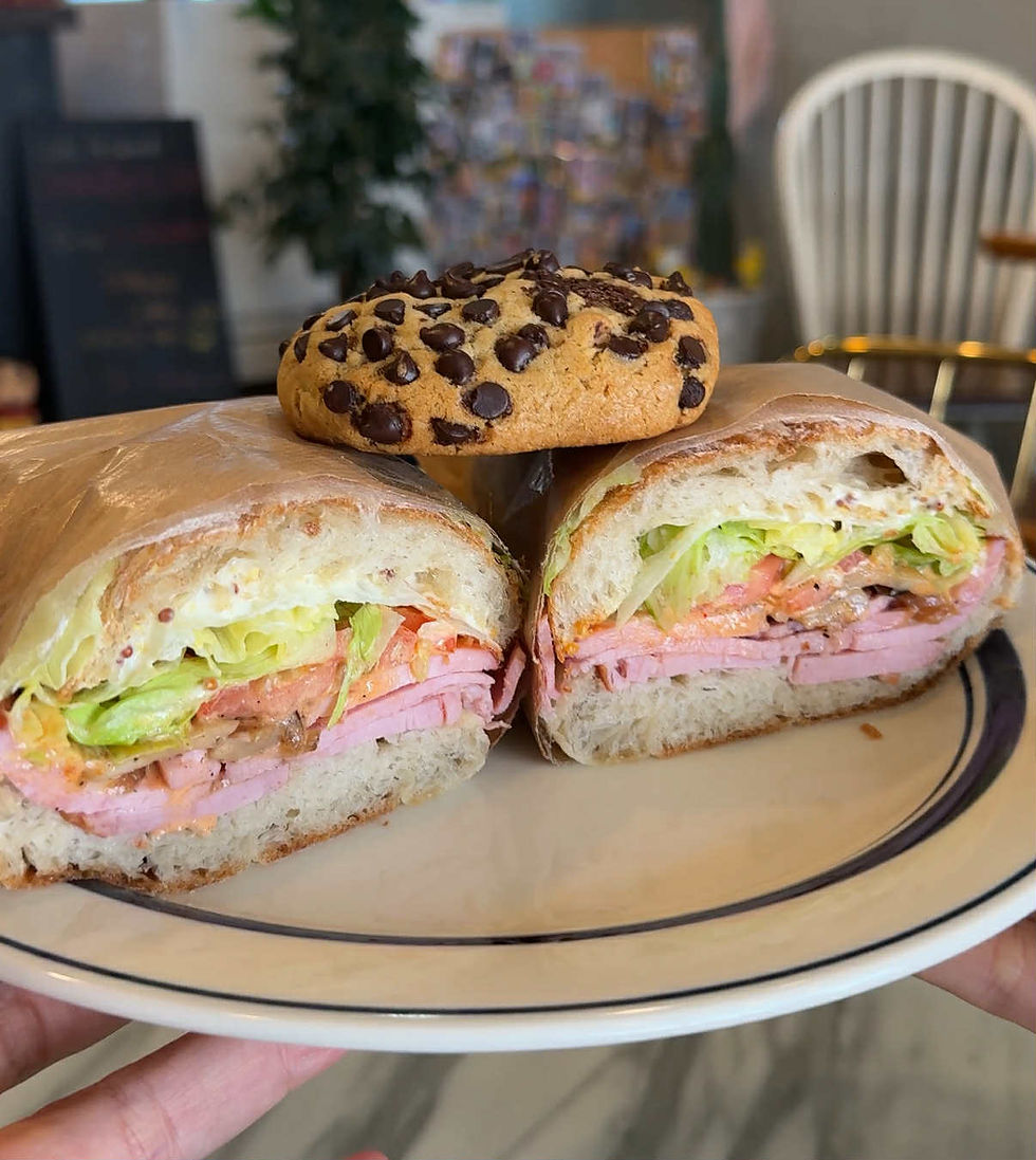 Plate with a ham and lettuce sandwich and a chocolate chip cookie. Background has a blurred plant and chair. Cozy cafe setting.