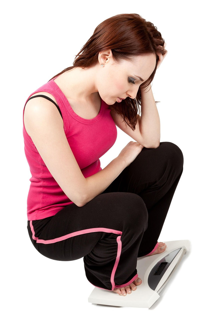 Woman in pink top and black pants crouched on a scale, hand on head, appearing thoughtful. White background, no text.
