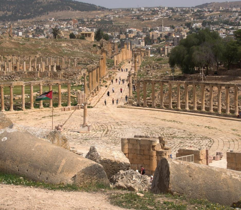 Ancient city with columns and flag, people walking on stone path. Background shows hills and modern buildings, clear sky, sunny day.