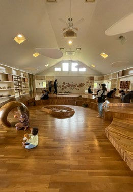 Spacious room with wooden floors and seating. People, including a child, relax and interact. Shelves and wall decor create a cozy vibe.