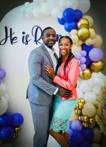 Couple smiling in front of a balloon arch with purple, white, and gold balloons. Text reads He is Risen. Joyful mood.
