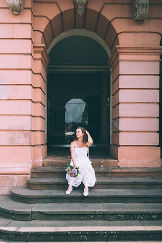Braut in Spitzenkleid sitzt mit Blumenstrauß und Sektglas auf einer Treppe vor einem historischen Gebäude – Hochzeitsfotografie von Fabio Bosco