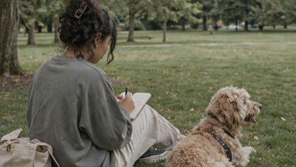 Woman writes in her journal on grass in a park, with a curly tan dog beside her and trees in the background.
