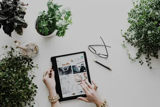 Manos usando tablet para diseño creativo rodeada de plantas y accesorios de escritorio.