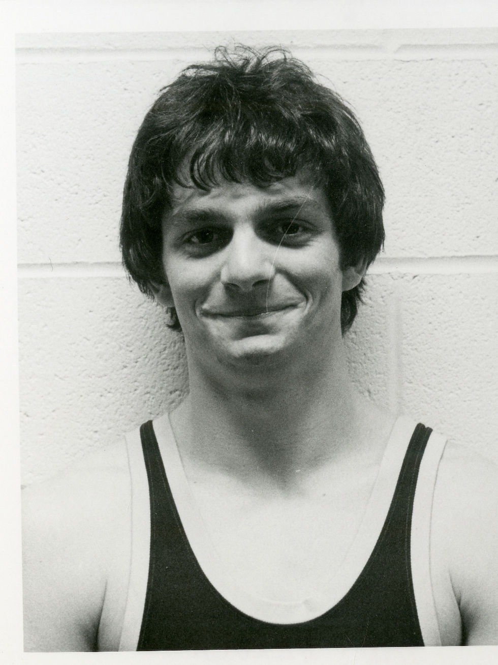 Young man in a dark sports tank top smiles slightly in a black and white photo. Plain brick wall background conveys a simple setting.