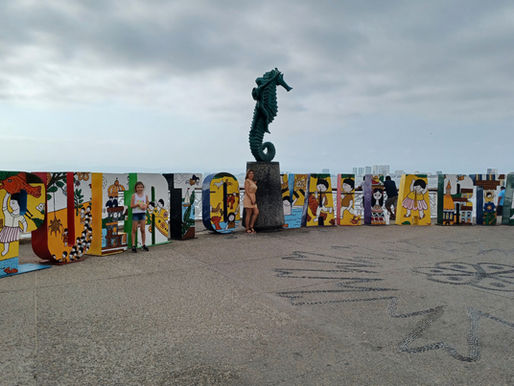 La mamá y la hermana de Yurani están de pie junto al colorido letrero de Malecón de Puerto Vallarta y la escultura del Caballito de Mar, con el océano y el perfil de la ciudad extendiéndose al fondo.